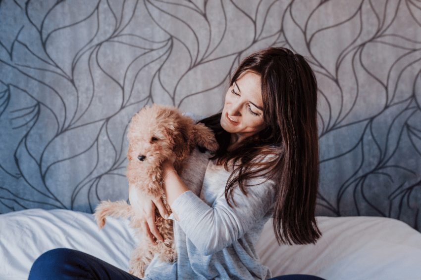 A woman with long dark hair is sitting on a bed, smiling affectionately at a small fluffy dog she is holding up. As a savvy pet owner, she's proud of her money-saving secrets. The background features a patterned wall with a leaf design.